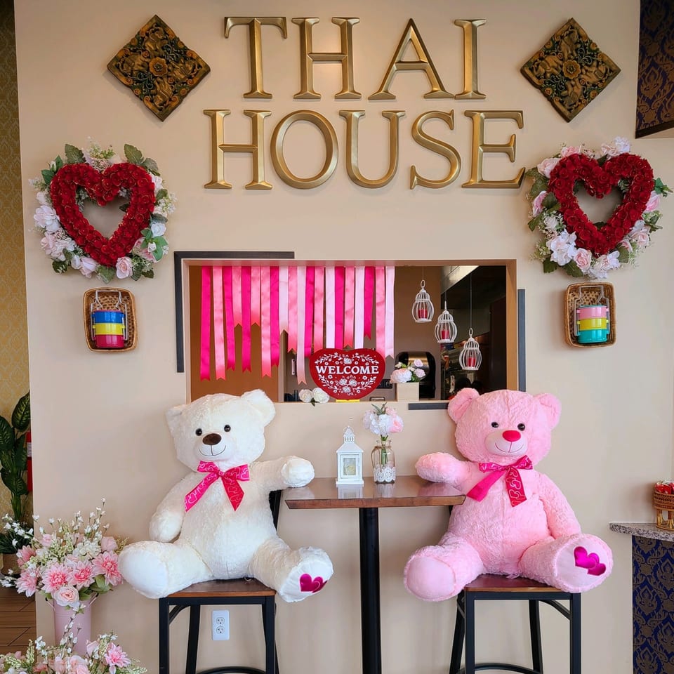 Two large teddy bears, one white and one pink, sit at a small table under a "THAI HOUSE" sign.