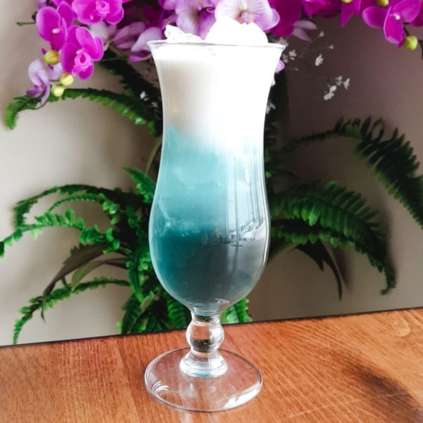A vibrant blue and white layered drink with ice, garnished with purple orchids and green ferns.