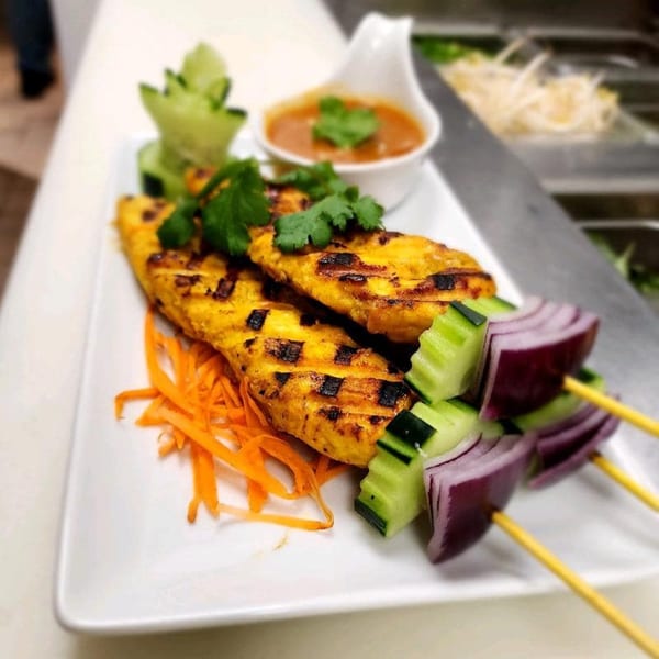 Grilled chicken satay with peanut sauce, shredded carrots, and vegetable skewers on a white plate.